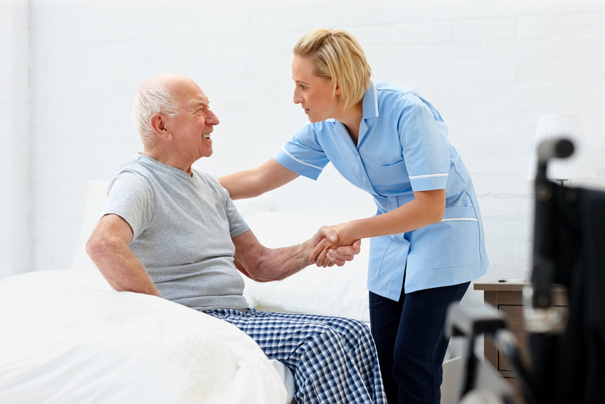Young female caregiver helping senior man get up from bed