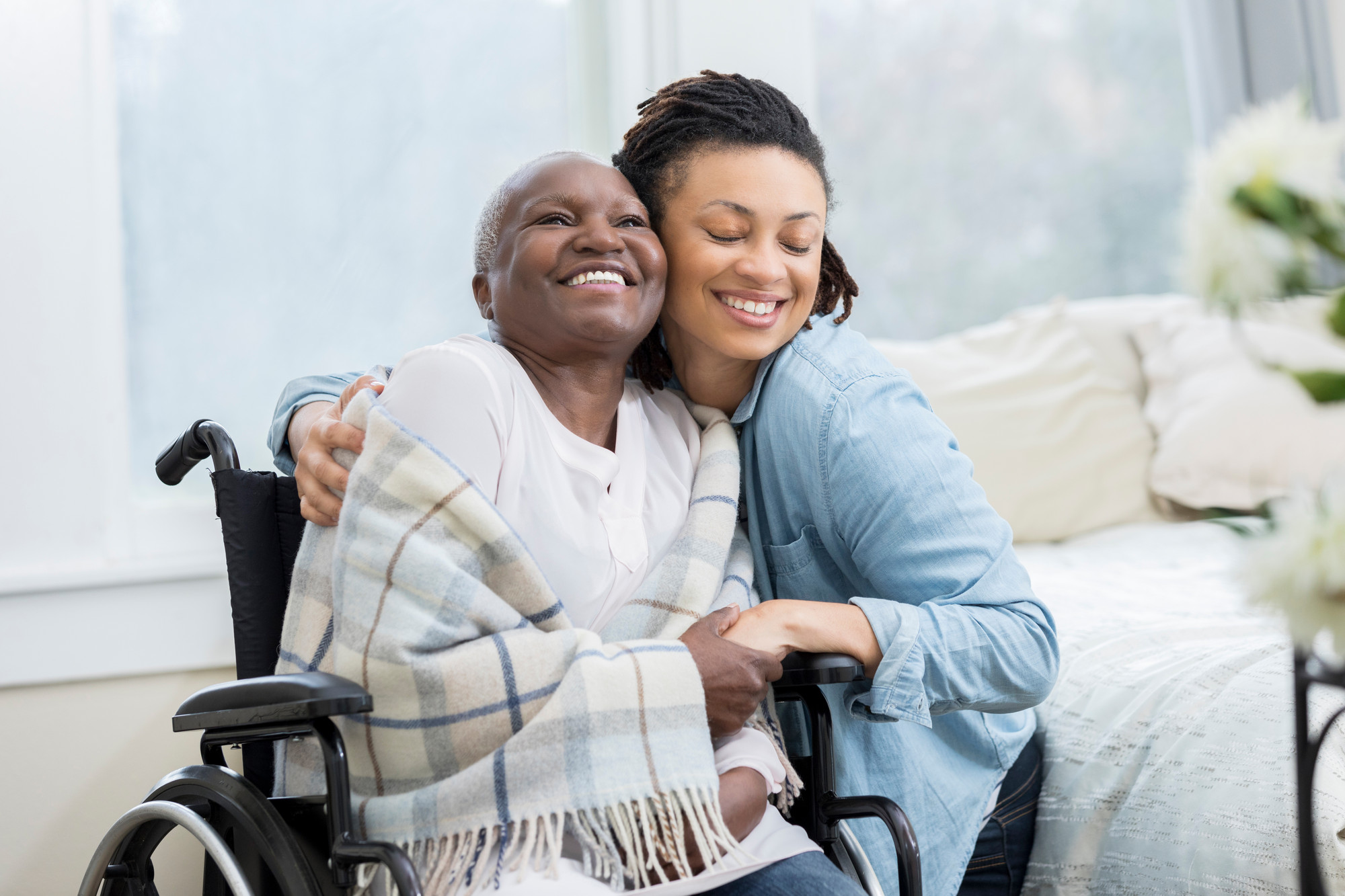 Young woman embraces her beautiful senior mother who is in a wheelchair. The adult daughter is visiting her mom in her mom's home.